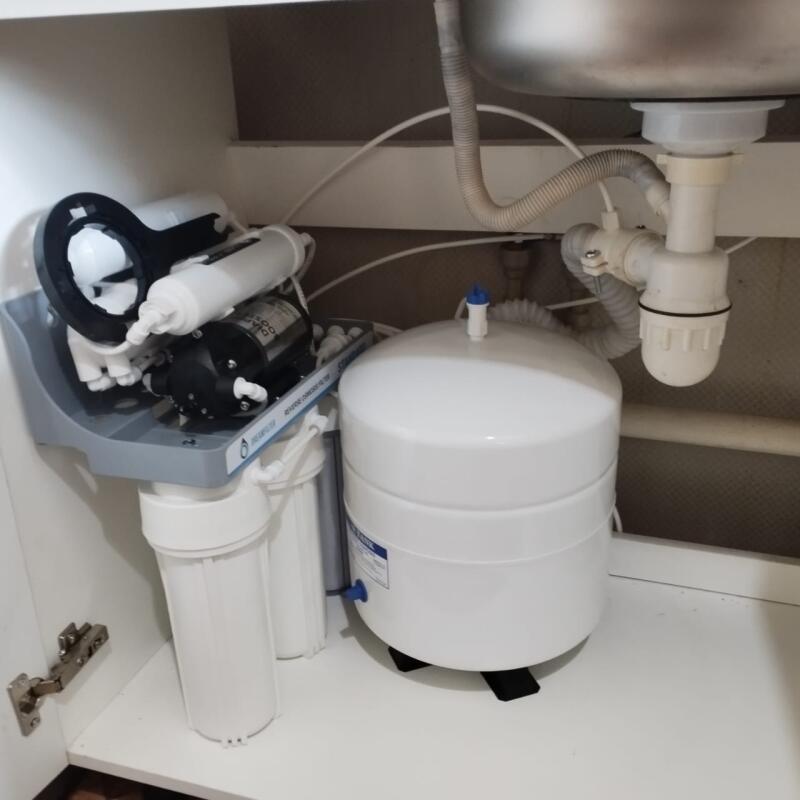 A reverse osmosis water filtration system with multiple filters, a pump, and a white storage tank is installed under a kitchen sink. Visible text includes "WATER FILTER" and "REVERSE OSMOSIS FILTER."