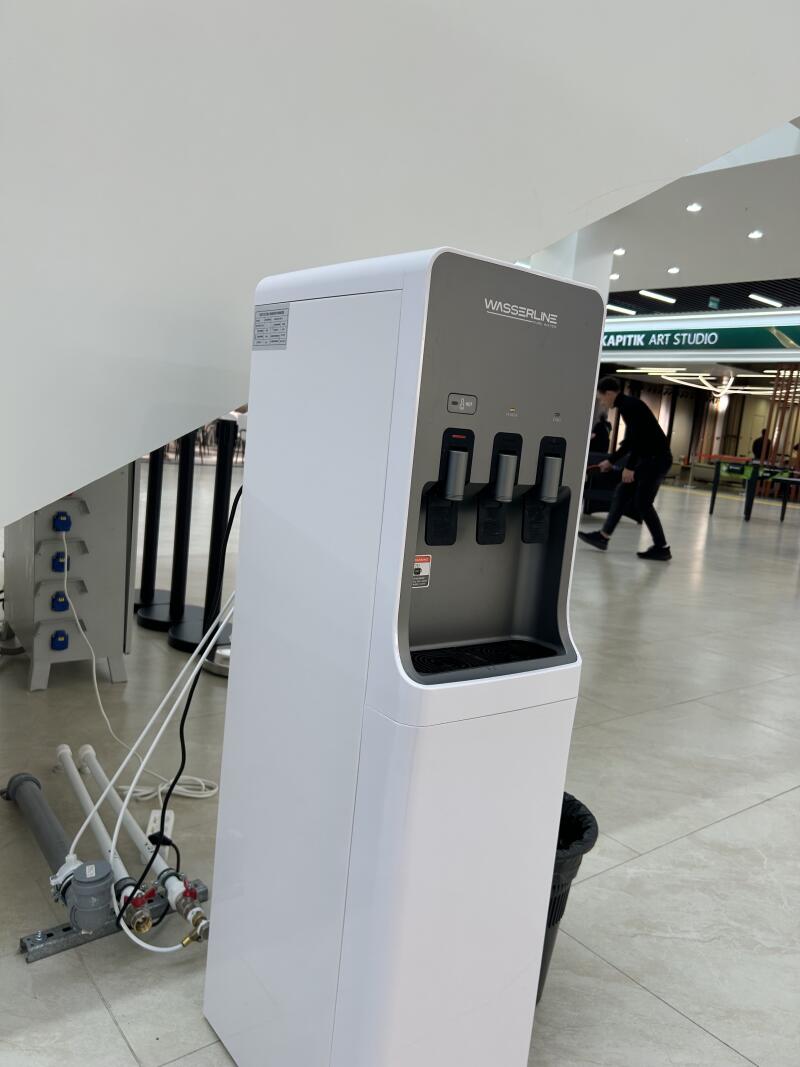 A white Wasserline water dispenser with three spouts stands next to a black trash can on a tiled floor, with a person and a "KAPITIK ART STUDIO" sign visible in the background.