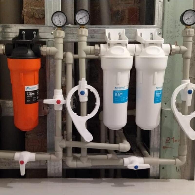 A plumbing system featuring one orange and two white water filters, branded "ecosoft" with pressure ratings, connected by light-colored pipes with pressure gauges and control valves.