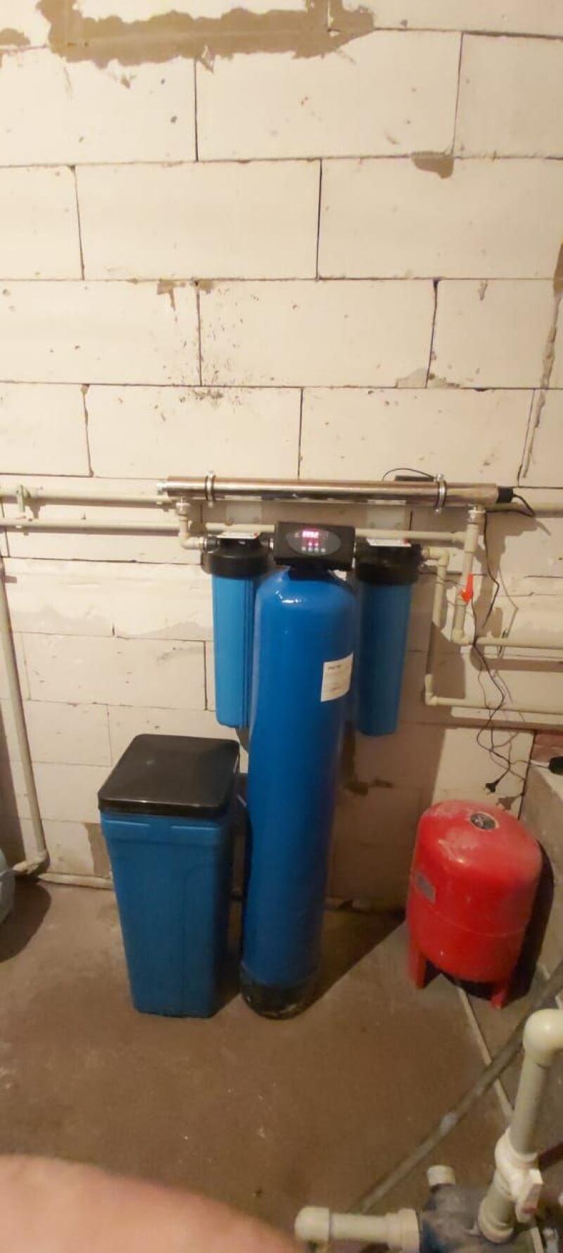 A blue water filtration system with a central tank, two smaller filters, and a digital control panel is installed against a white brick wall, with a tall blue container with a black lid to its left and a red pressure tank on the concrete floor to its right.