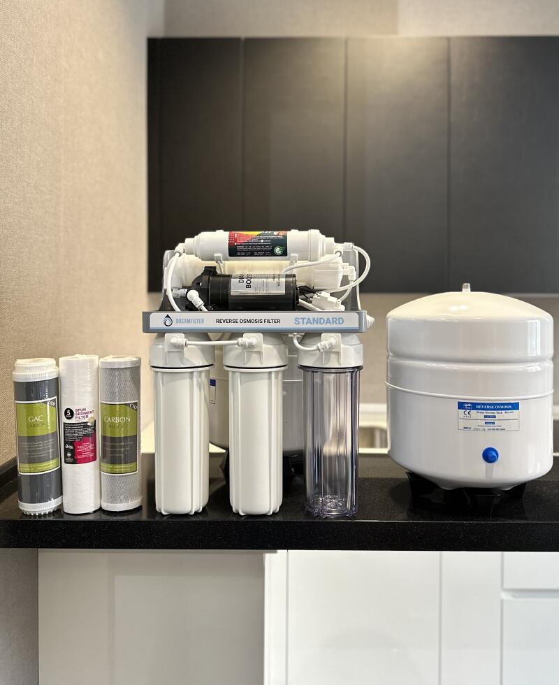 A reverse osmosis water filtration system, including multiple filters, a separate pressure tank, and three additional filter cartridges (GAC CARBON, SPUN SEDIMENT FILTER, CARBON), is displayed on a black countertop against a dark grey cabinet background. The main system is labeled "DREAMFILTER REVERSE OSMOSIS FILTER STANDARD."