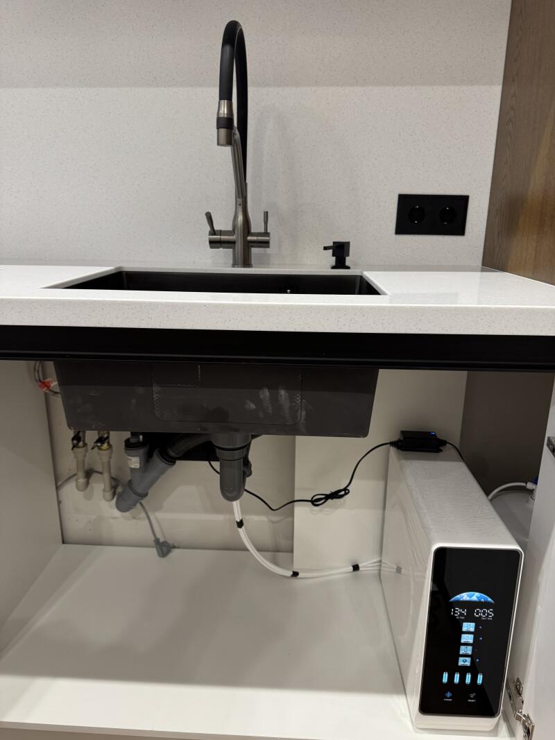 A kitchen sink setup featuring a dark grey sink, a brushed metal faucet with a black flexible hose, and a black soap dispenser on a white speckled countertop and backsplash, with black power outlets. Underneath, visible plumbing connects to a white water filtration system with a digital display showing "134 005" and various icons.