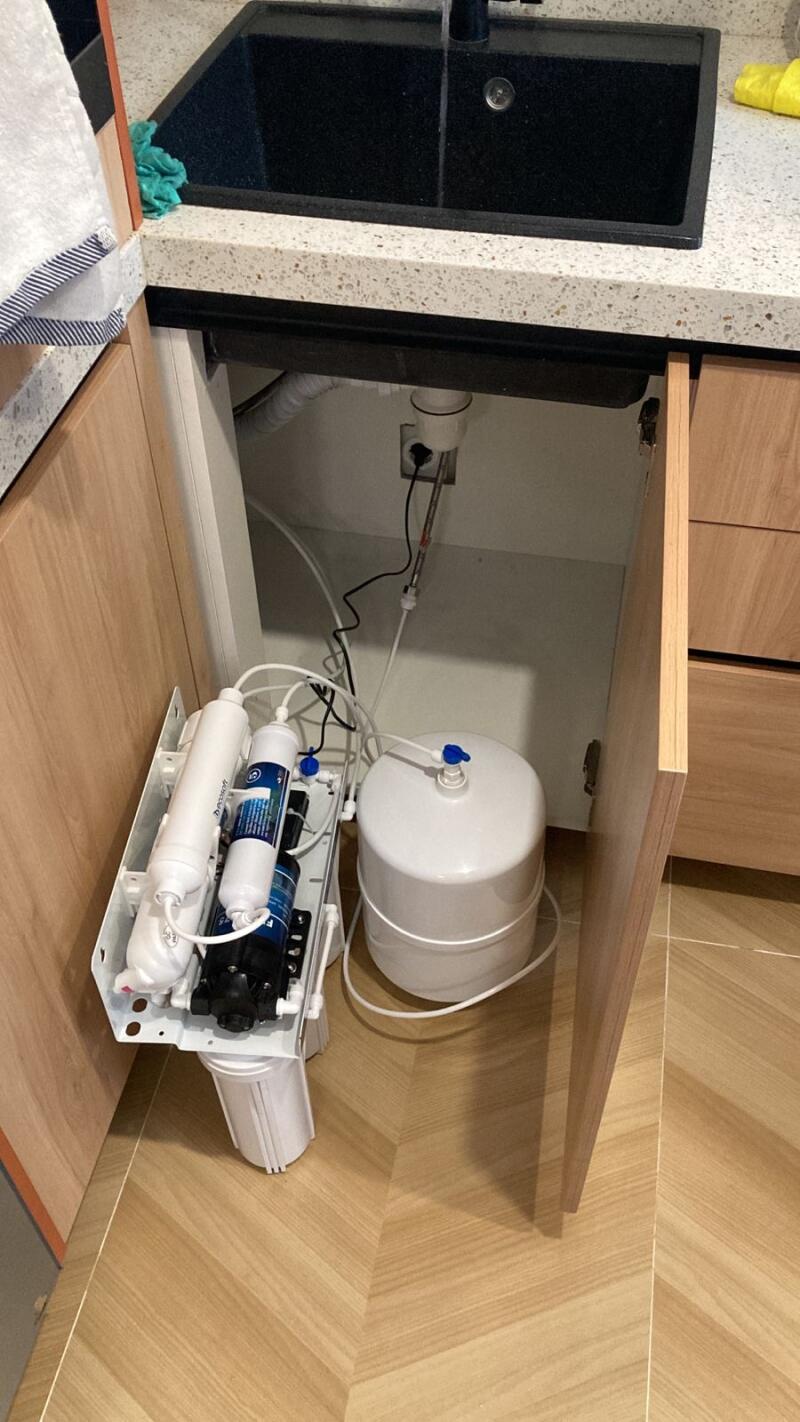 A water filtration system with multiple filter cartridges and a pressure tank is visible on the floor and inside an open cabinet under a black kitchen sink with a speckled white countertop.