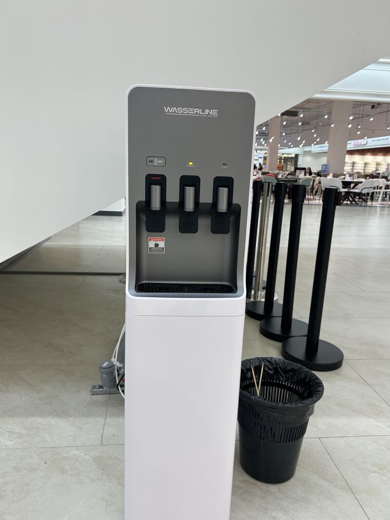 A white and grey WASSERLINE water dispenser with three spigots and a yellow power indicator light stands next to a black trash can and queue stanchions on a tiled floor in a bright indoor area.