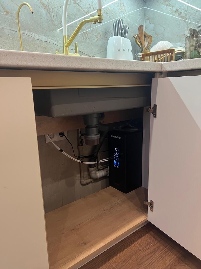 A kitchen sink area with a gold faucet and a white Smeg knife block, with the cabinet underneath open to reveal plumbing and a black 'Dreamfilter' water purification system displaying '89'.