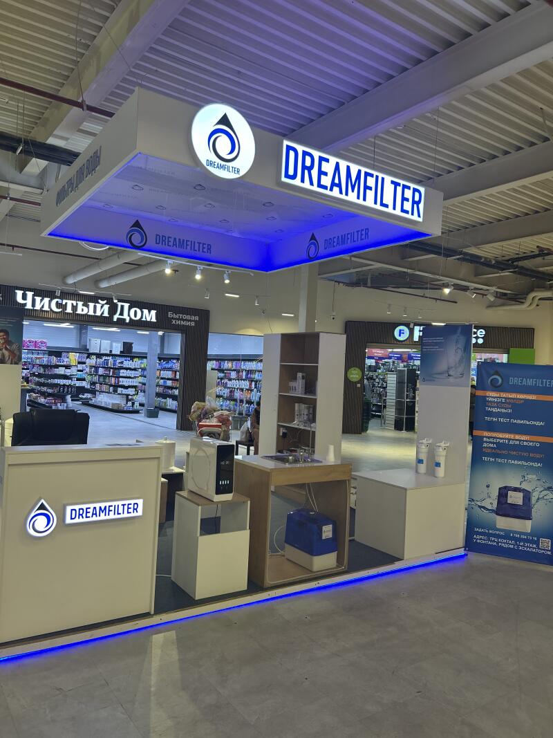 A brightly lit 'DREAMFILTER' water filter stand inside a store, featuring a prominent hanging sign with the company name and logo, blue LED lighting, various filtration units, and a promotional banner with text in Kazakh and Russian. In the background, another store sign reads 'ЧИСТЫЙ ДОМ' and 'Бытовая химия'.
