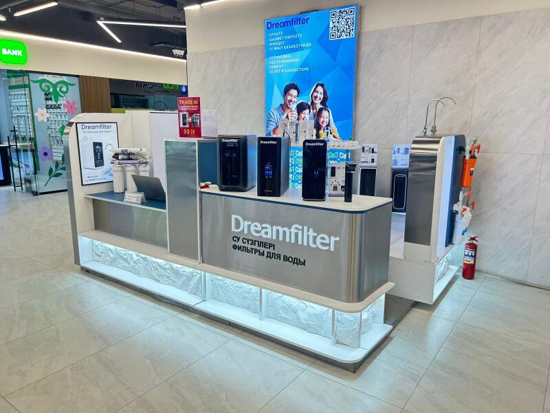A Dreamfilter brand stand in a store displays various water filter models, a laptop, and a large blue banner featuring a family, a QR code, and the text "Dreamfilter СУ СҮЗГІЛЕРІ ФИЛЬТРЫ ДЛЯ ВОДЫ". A "TRADE IN" sign and a red fire extinguisher are also visible.