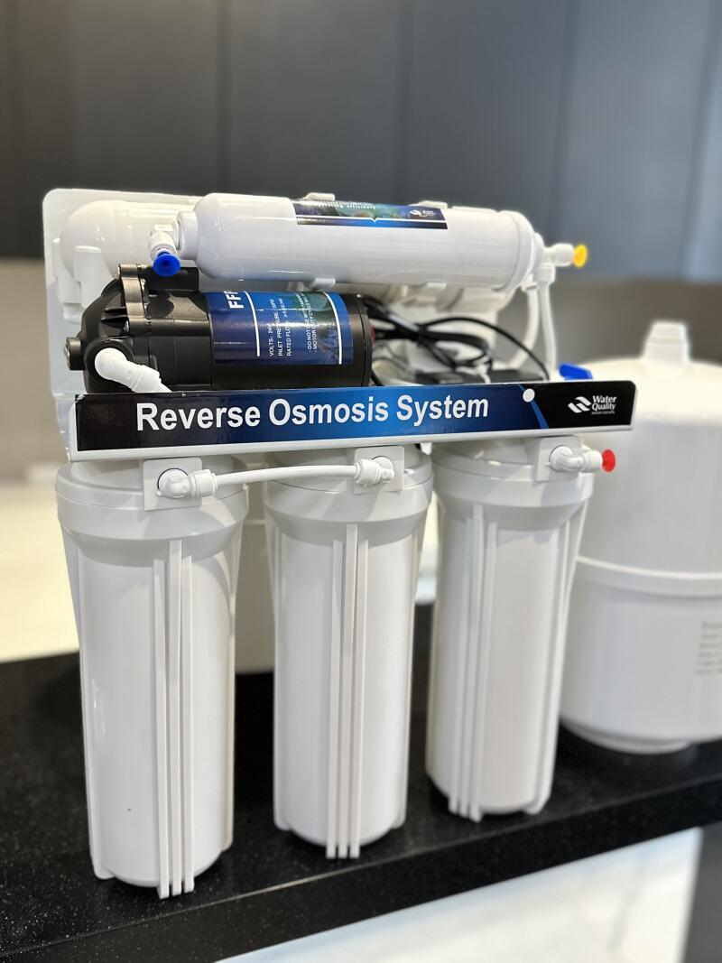 A white reverse osmosis water filtration system featuring three large filter housings, a pump, and an upper filter cartridge, with a blue banner displaying 'Reverse Osmosis System' and a 'Water Quality' logo.