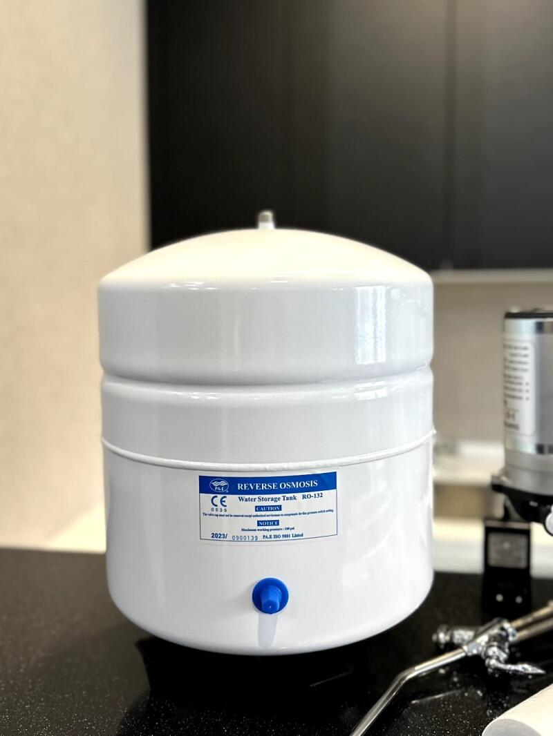 A white reverse osmosis water storage tank with a blue label displaying "REVERSE OSMOSIS", "Water Storage Tank RO-132", "CE 0035", and other specifications, sits on a dark surface.