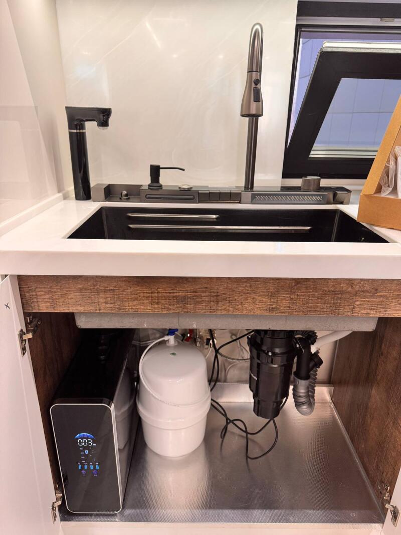 A modern kitchen sink setup with two faucets, a soap dispenser, and a window in the background. Below the sink, inside the cabinet, are a black water filter system displaying "003-", a white water tank, and a garbage disposal unit.