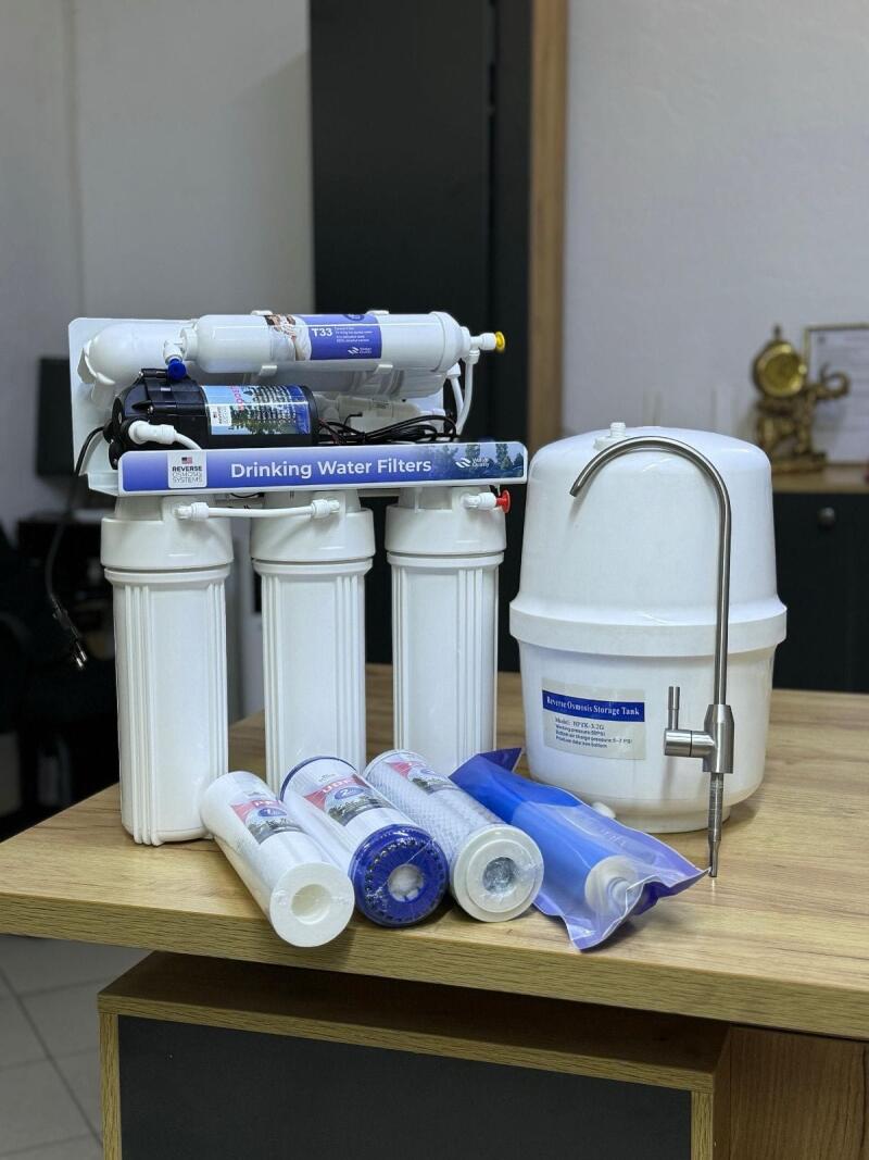 A water filtration system with multiple white filter housings, a blue pump, and filters labeled "T33" and "Drinking Water Filters" is displayed next to a white reverse osmosis storage tank with a chrome faucet and a "Reverse Osmosis Storage Tank" label. Several new filter cartridges, some in blue packaging, are on the wooden table in front.