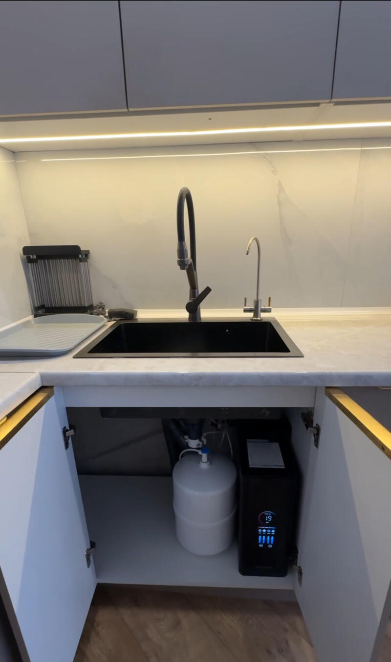 A modern kitchen sink area features a black sink, two faucets, and a white marble countertop under strip lighting. To the left is a dish drying rack, and under the sink, a white water tank and a black water filter system with a digital display showing '19' are visible.