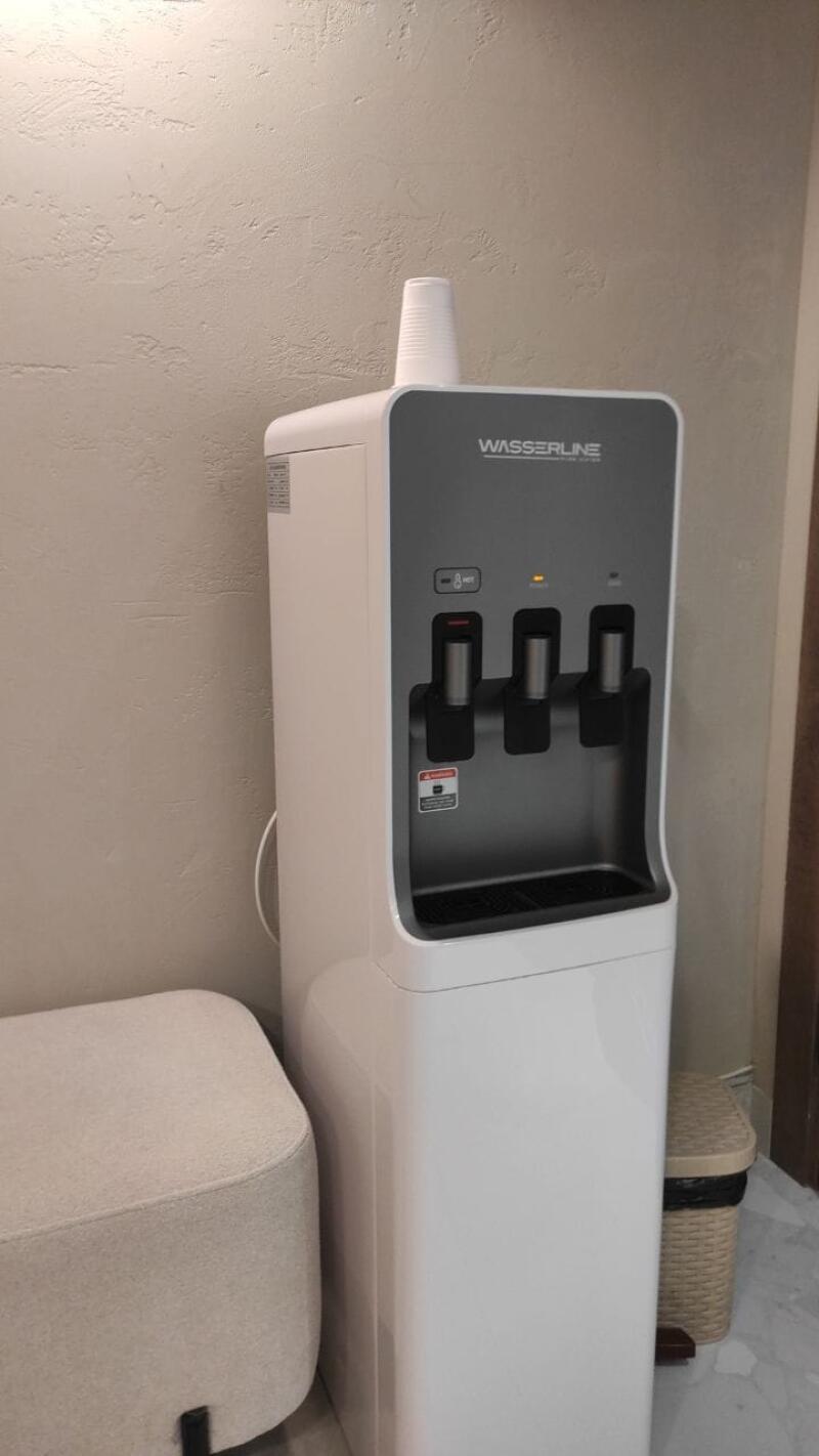 A white and grey WASSERLINE water cooler with three dispensers stands against a textured light wall, with a white paper cup on top, next to a light-colored ottoman and a woven trash can.