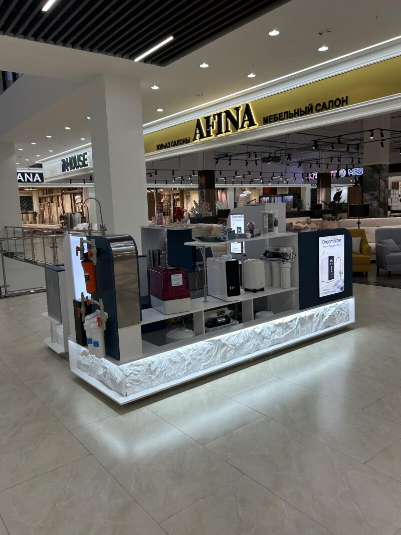 A white display stand showcasing water filtration systems and related appliances is visible in a mall, with background shop signs reading "AFINA", "МЕБЕЛЬНЫЙ САЛОН" (Furniture Salon), "Dreamfilter", "INHOUSE", and "ANA".