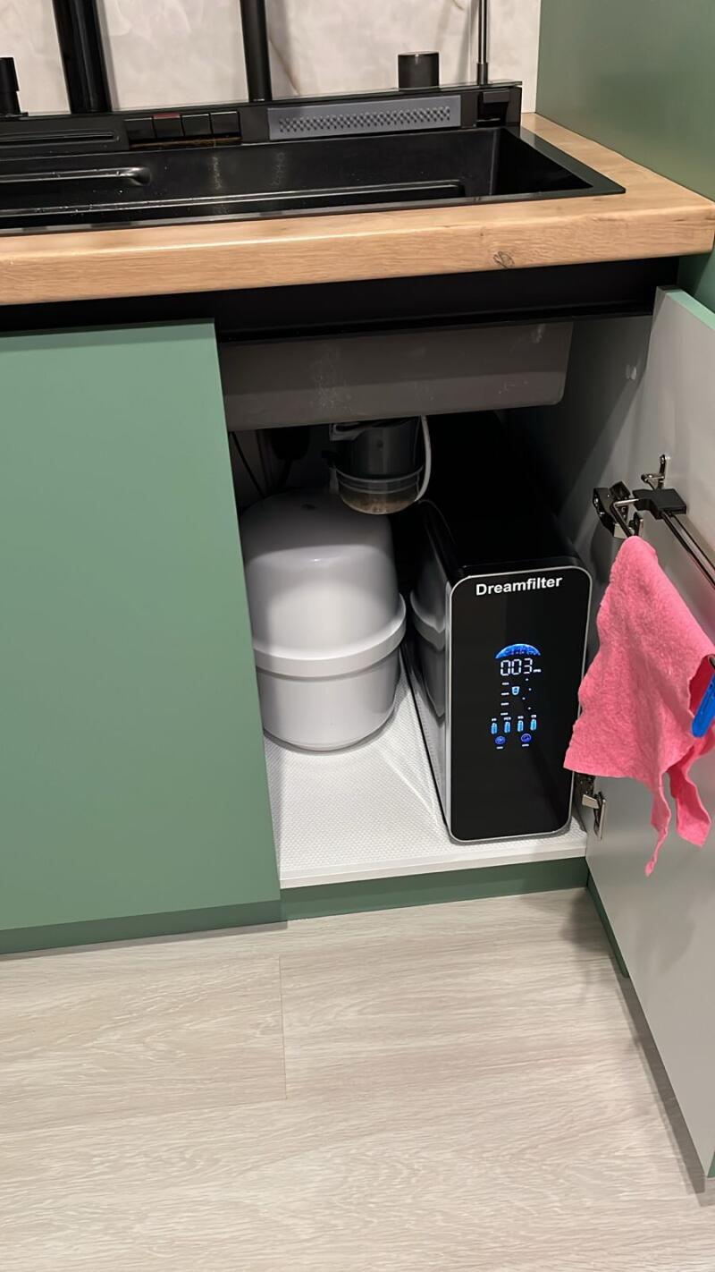 An open under-sink cabinet features a white cylindrical water tank and a black 'Dreamfilter' water filter with a digital display showing '003-', while a pink cloth hangs on the inside of the cabinet door below a black sink and wooden countertop.