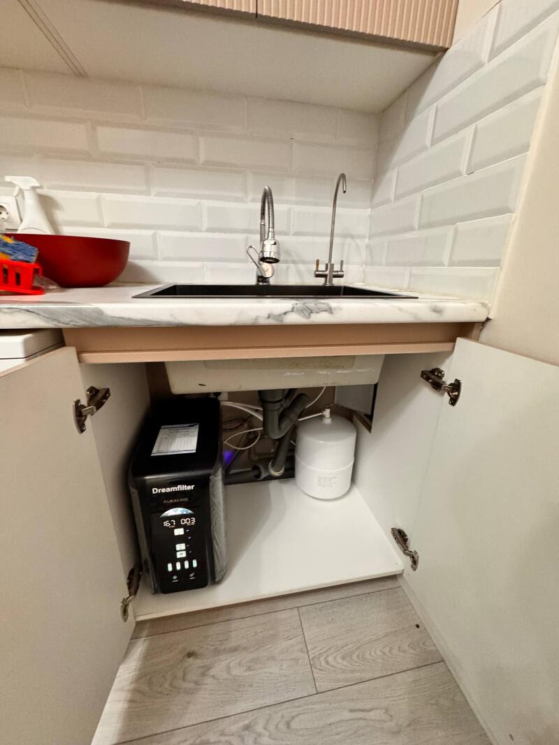 A kitchen sink area with white subway tiles, a black sink, and a marble-patterned countertop. Underneath, with open cabinet doors, are a black "Dreamfilter ALKALINE" water filter system with a digital display "16.1 003" and a white cylindrical tank.