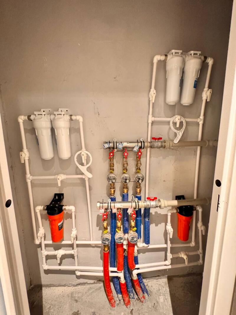 A complex plumbing system with white pipes, several water filters (white and orange), and manifolds with red and blue insulated pipes, pressure gauges, and red valves, installed on a gray wall.