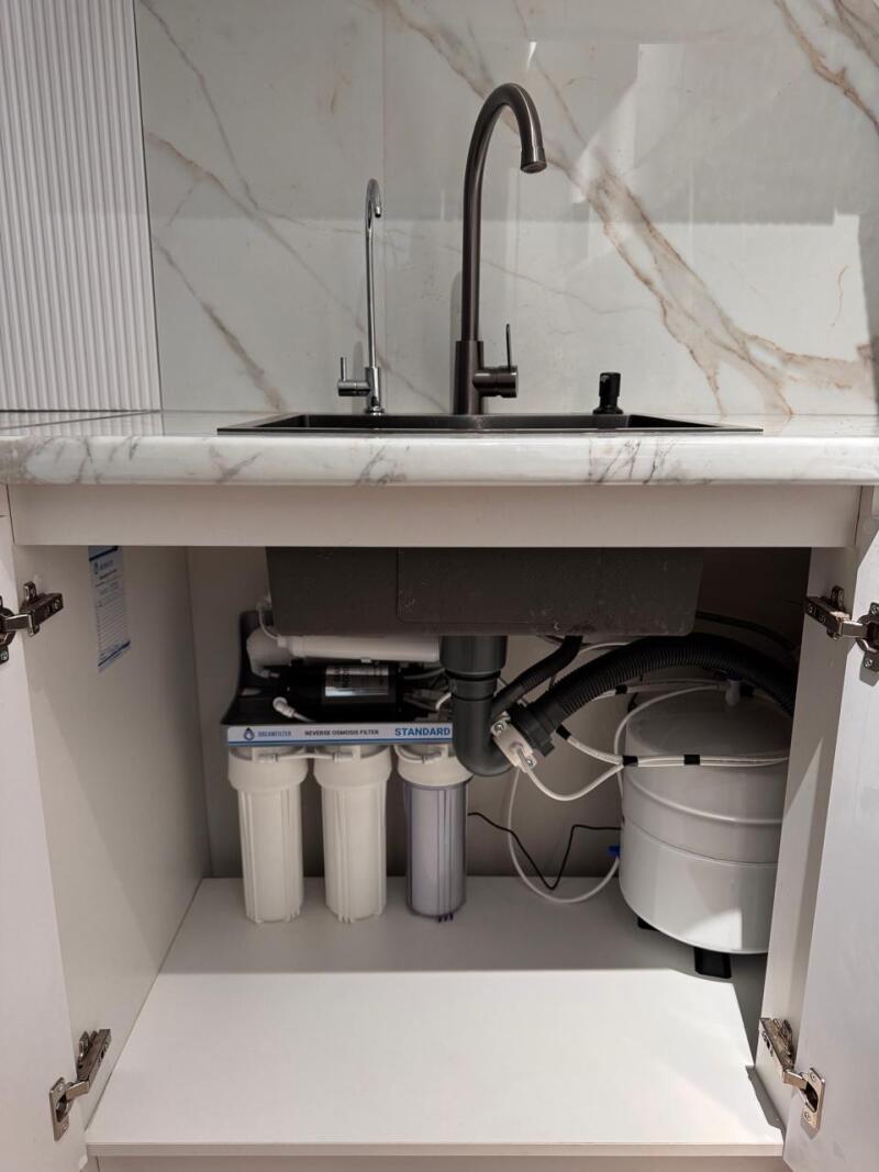 A kitchen sink area features a black sink with two faucets, one dark and one silver, set against a white marble-patterned countertop and backsplash. Below the sink, inside an open white cabinet, a water filtration system labeled 'REVERSE OSMOSIS FILTER STANDARD' includes three filter housings and a white storage tank.