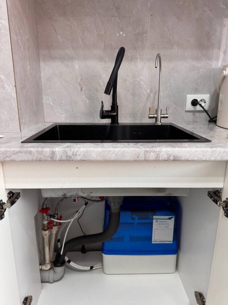 A black kitchen sink with a black faucet and a separate silver water filter faucet is set in a gray marble-patterned countertop. Below the sink, an open cabinet reveals plumbing pipes with red valves and a blue and white water filtration system.