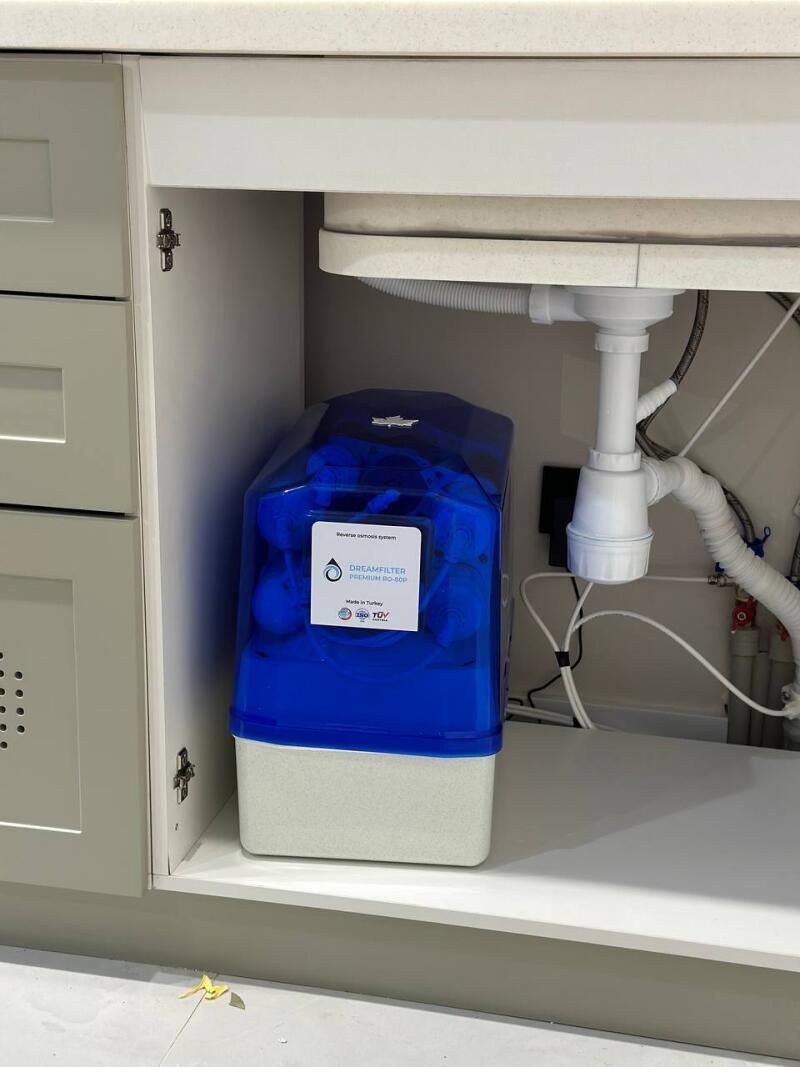 A blue and white Dreamfilter Premium RO-50P reverse osmosis water filter system, labeled 'Made in Turkey', is placed under a sink in a light grey cabinet, with pipes and hoses visible.