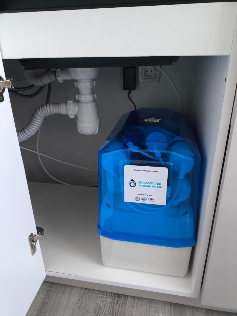 A blue and white Dreamfilter Premium RO-80P reverse osmosis system is placed inside a white cabinet under a sink, with plumbing pipes, an electrical outlet, and an open cabinet door visible.