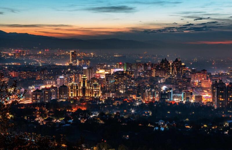 A panoramic view of a sprawling city at twilight, featuring numerous illuminated buildings and a few mountains in the background under a colorful sky.