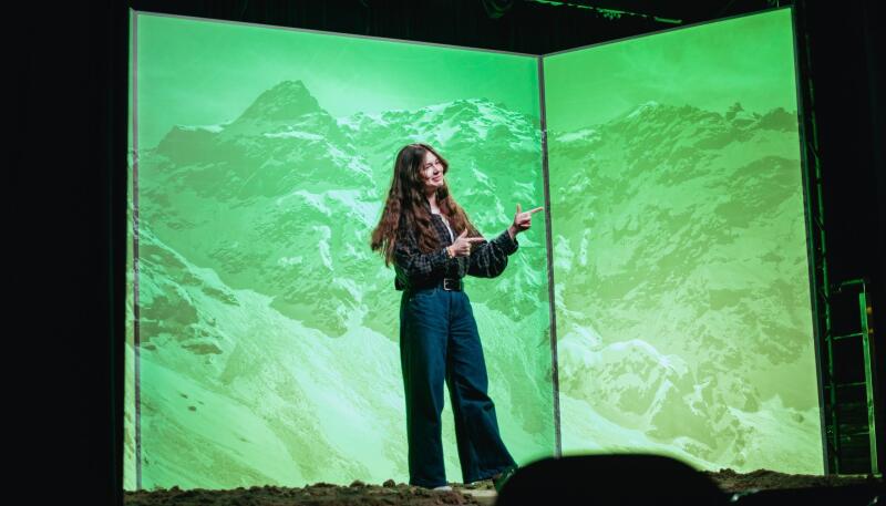 A young woman with long brown hair, wearing a plaid shirt and dark jeans, stands on a dark textured surface, pointing with both hands at two large screens behind her that display a green-toned image of snowy mountains.