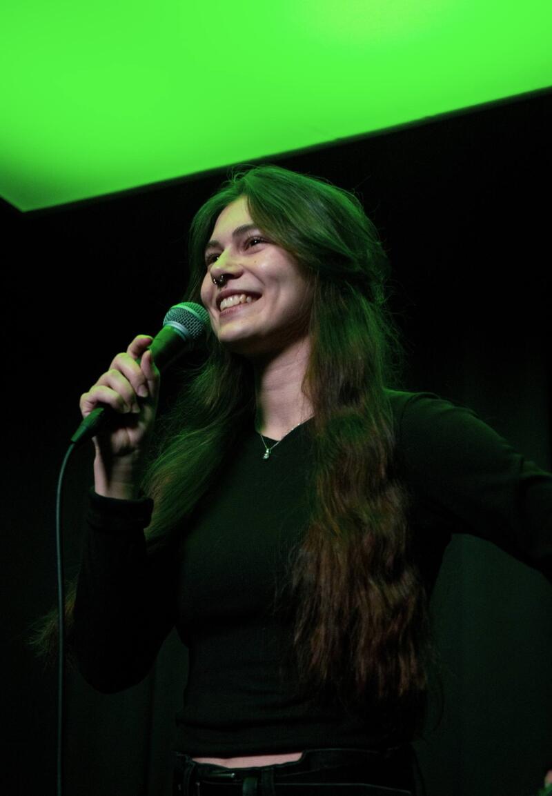 A smiling young woman with long dark hair and a nose ring holds a microphone, illuminated by a green light from above against a dark background.