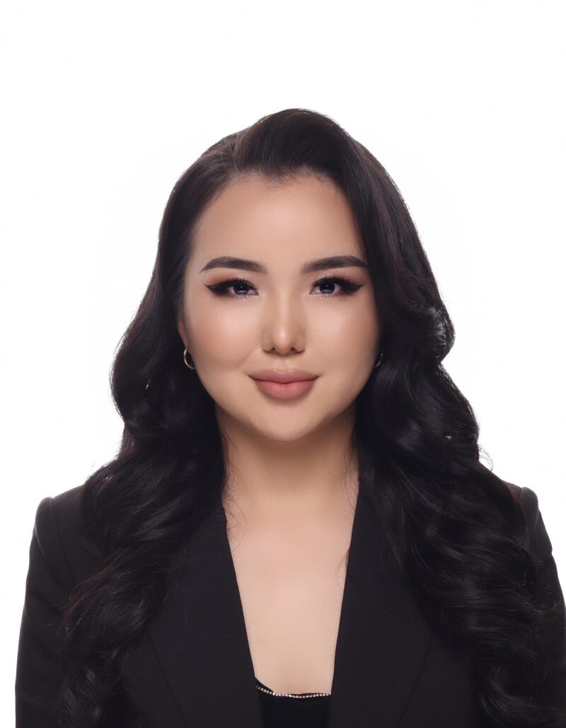 A woman with long dark wavy hair and makeup, wearing a black blazer and small hoop earrings, looks directly at the camera against a white background.