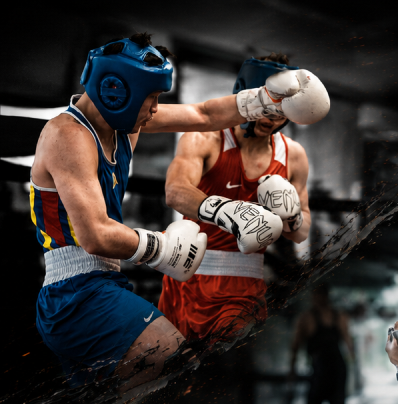 Two boxers wearing protective headgear and white gloves are actively sparring in a ring; the boxer in blue with "IBS" on his glove is throwing a punch towards the boxer in red, whose gloves read "VENUM".