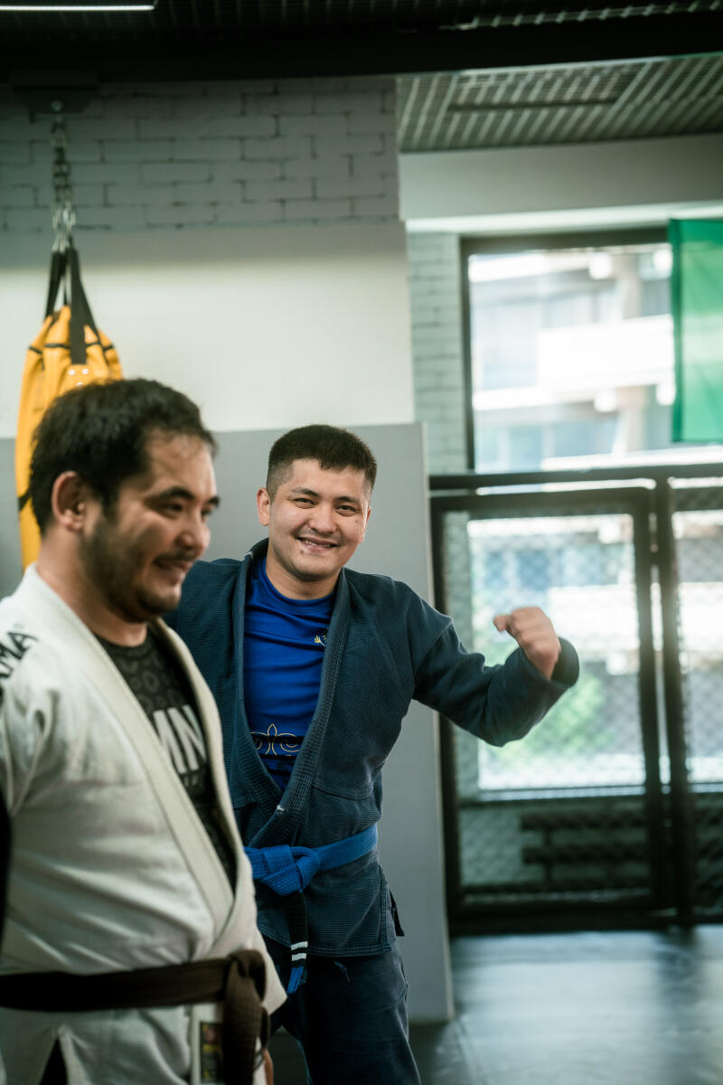 Два мужчины в кимоно для боевых искусств находятся в спортзале; один в синем кимоно с синим поясом улыбается и сгибает руку, а другой в белом кимоно с коричневым поясом частично виден слева.