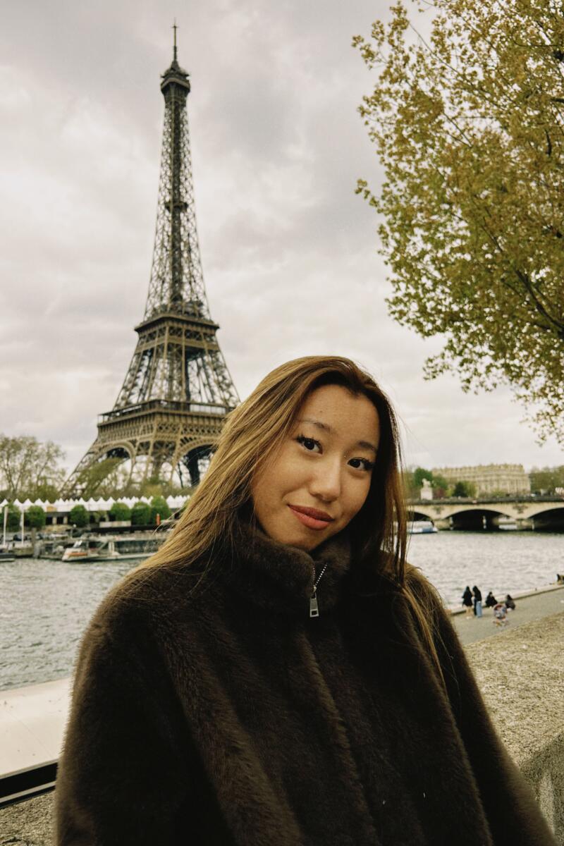 A woman with long brown hair and a dark fur jacket smiles at the camera, with the Eiffel Tower, the Seine River, a bridge, and trees under a cloudy sky in the background.