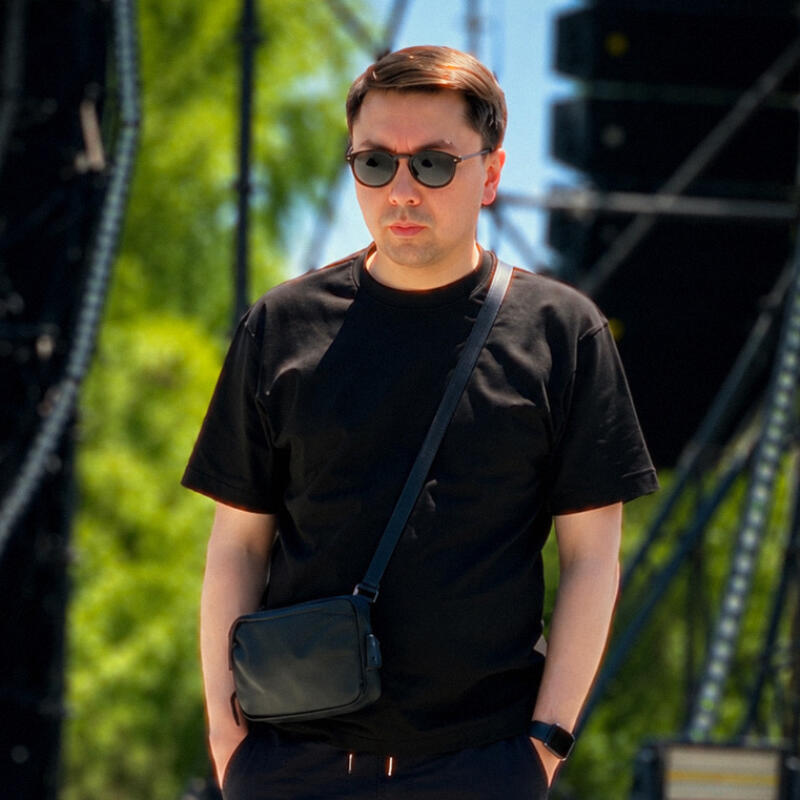 A man wearing a black t-shirt, black pants, sunglasses, and a black cross-body bag stands outdoors with hands in pockets, against a blurred background of greenery and stage structures.