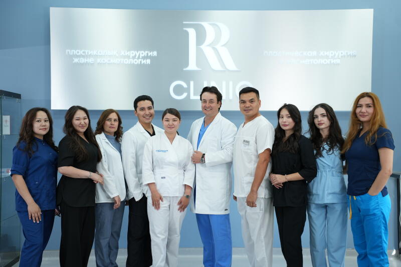 A group of nine medical professionals, including seven women and two men, stands in a clinic with a sign in the background that reads "RR CLINIC," "пластикалық хирургия және косметология," and "пластическая хирургия и косметология."