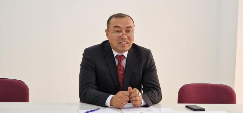 A man in a black suit, white shirt, and red tie sits at a table with papers, a pen, and a smartphone, his hands clasped. He wears glasses and appears to be speaking.
