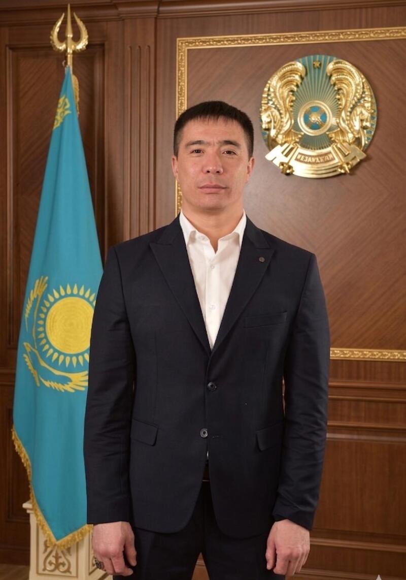 A man in a dark suit and white shirt stands facing forward, with the blue and gold flag of Kazakhstan to his left and the golden national emblem of Kazakhstan with the text "QAZAQSTAN" to his right, against a wooden wall.