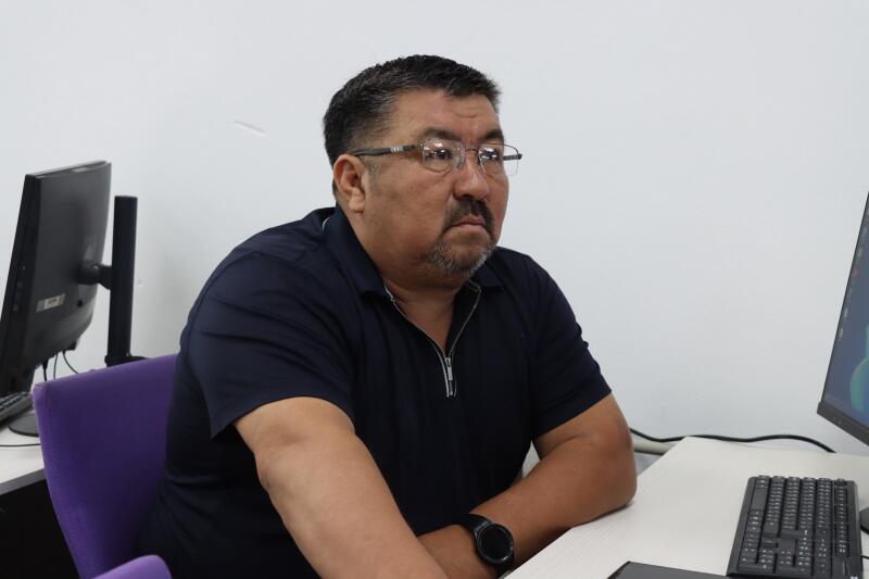 A man with glasses, a beard, and a dark blue polo shirt sits at a desk with computer equipment, looking to the right.