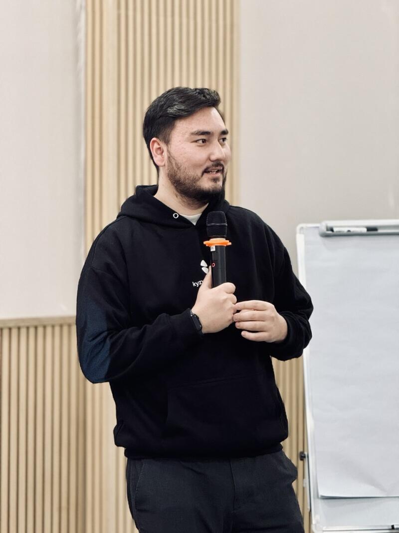 A smiling man with a beard and dark hair, wearing a black hoodie with 'kyz' text and black pants, holds a microphone with an orange ring. He stands in front of a light wall with vertical wooden slats and a white flip chart.