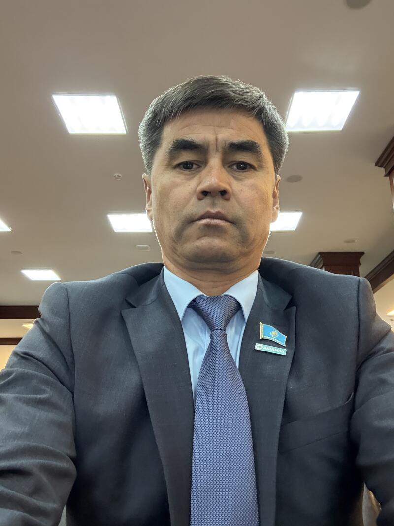 A man in a dark suit, light blue shirt, and blue patterned tie looks directly at the camera. He wears a pin of the Kazakhstan flag and a pin with "AMANAT" written on it on his lapel.