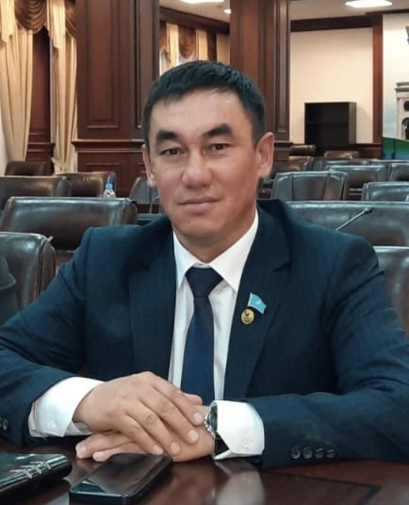 A man in a dark suit, white shirt, and blue tie with a Kazakhstan flag pin on his lapel sits at a wooden table with clasped hands, looking at the camera. Two smartphones are on the table, and a conference room with leather chairs is in the background.
