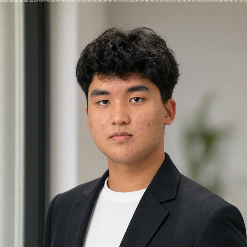 A young Asian man with black curly hair, wearing a white t-shirt and black blazer, looks directly at the camera with a neutral expression against a blurred light background.