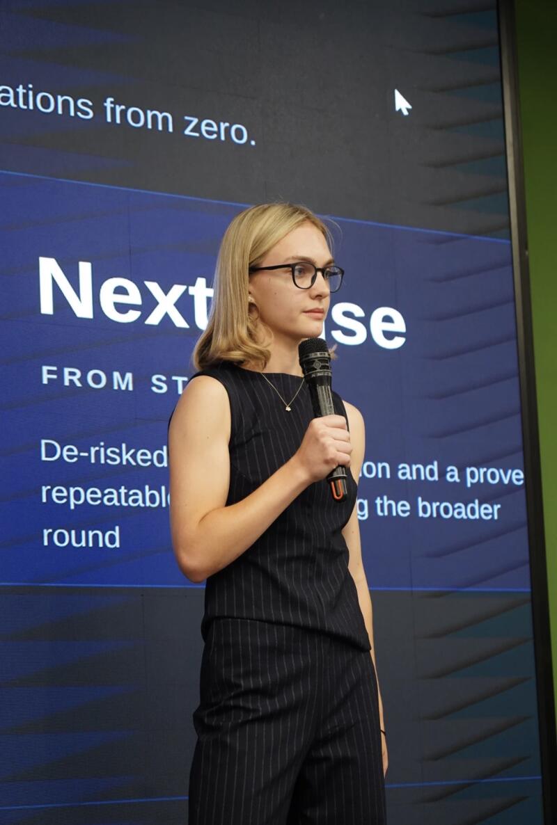 A young woman with blonde hair and glasses, wearing a dark pinstriped sleeveless top and matching pants, holds a microphone and stands in front of a blue screen displaying text such as "Next phase", "De-risked repeatable", and "from zero".