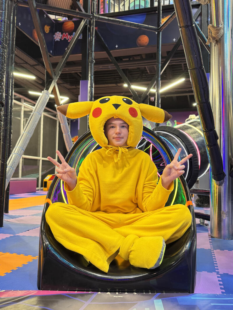 A person wearing a yellow Pikachu costume sits cross-legged in a black curved structure, making peace signs with both hands. The background shows an indoor play area with space-themed elements, including planets and "#My!" text, and colorful floor mats.