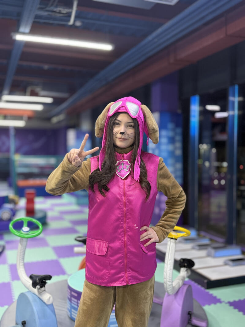 A young person in a Skye from Paw Patrol costume, consisting of a pink vest, brown long-sleeved top and pants, and a pink hat with goggles and ears. The person has whiskers painted on their face, makes a peace sign with one hand, and stands in an indoor play area with a checkered floor.