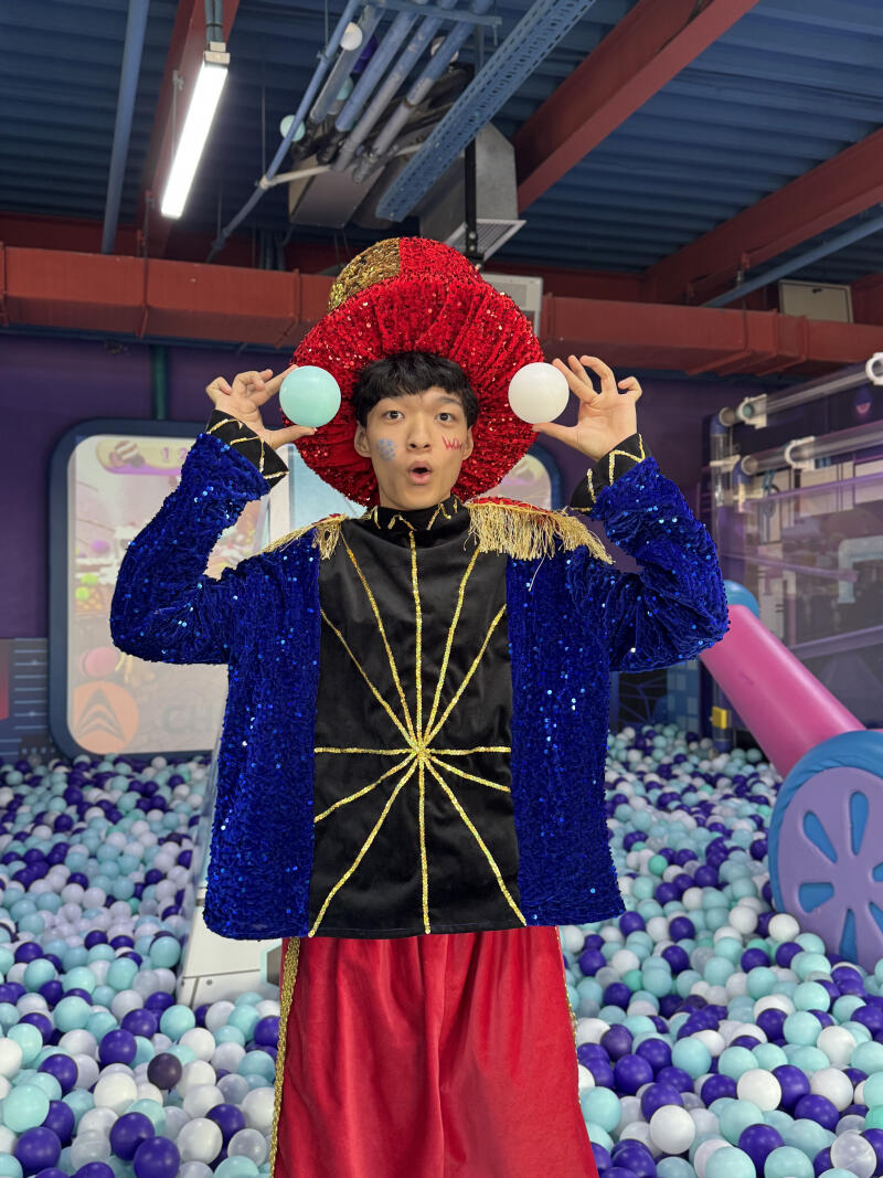 A person in a red sequined hat and blue sequined jacket stands in a ball pit, holding a light blue and a white ball near their head. They have a surprised expression and face paint, including a red 'W' on their cheek.