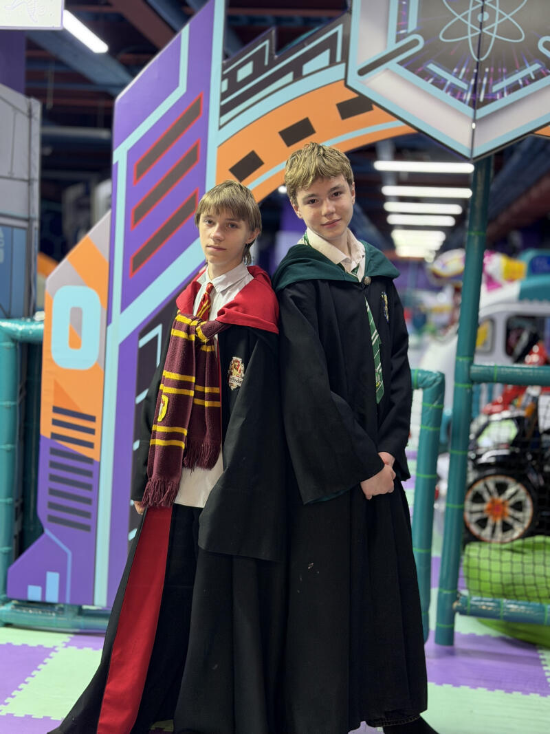 Two boys dressed in Harry Potter-themed robes and scarves stand in an indoor play area with colorful abstract decorations. One boy wears a red Gryffindor scarf, and the other wears a green Slytherin tie.