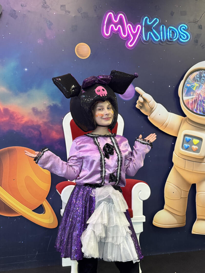 A person in a purple costume and a black Kuromi hat with a pink skull sits on a chair against a space-themed backdrop with planets and an astronaut, under a neon sign that reads "My Kids". The person has "WOW" written on their cheek.