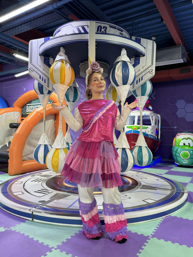 A woman in a vibrant pink and purple unicorn costume stands on a circular platform in an indoor amusement park, surrounded by colorful hanging decorations and play equipment, with "WOW" written on her cheek and "03" visible on a structure above.