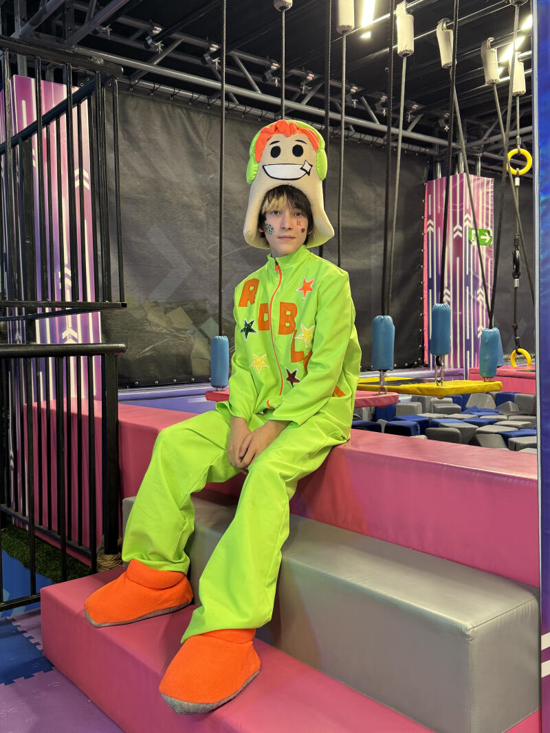 A young person sits on pink and grey padded steps in an indoor play area, wearing a neon green tracksuit with orange 'ROBLY' text and stars, an orange and green character hat, and orange slippers.