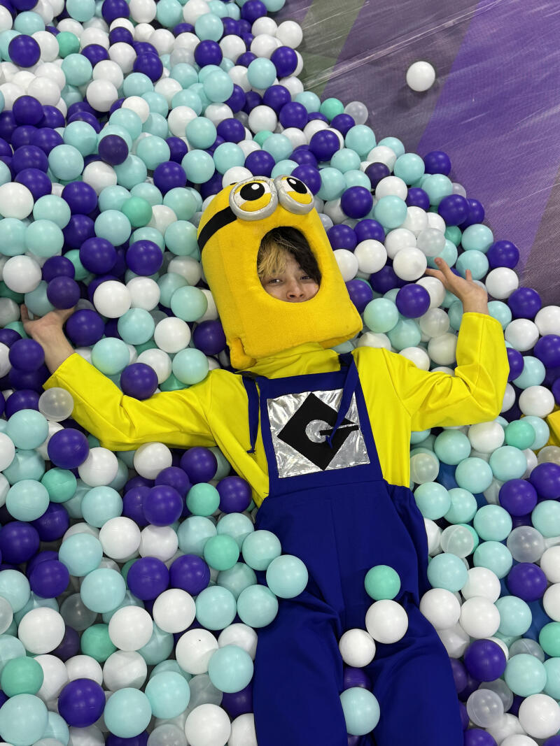 A person dressed in a yellow Minion costume with blue overalls and a Gru logo is lying in a ball pit filled with white, light blue, and purple balls.